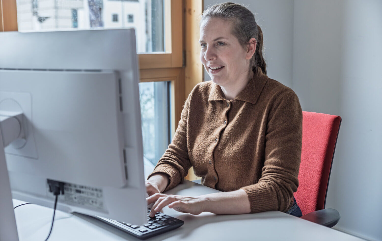 Eine junge Frau mit braunem Pullover sitzt im Büro an ihrem Schreibtisch.