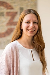 Eine Frau mit braunen langen Haaren mit einem weißen Oberteil und einer pinken Strickjacke.