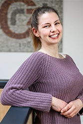 Mitarbeiterin im Portrait. Mitarbeiterin in fliederfarbenen Pullover lehnt an einer hölzernen Ballustrade.