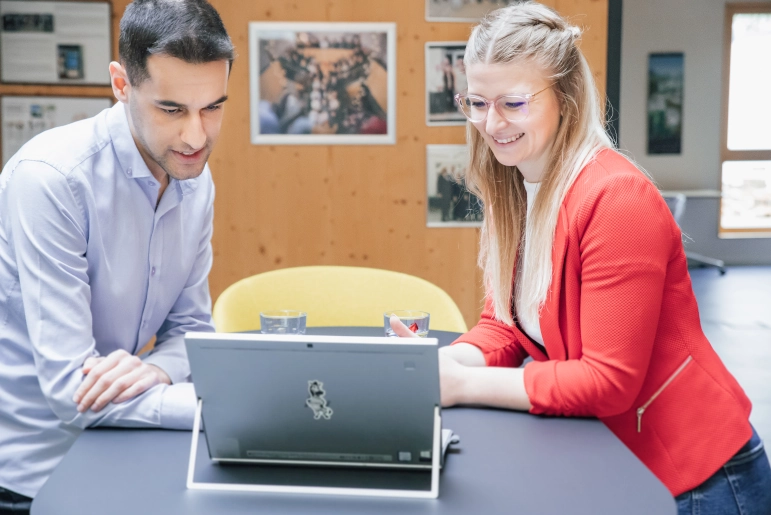 Eine Frau in rotem Blazer und blonden Haaren steht an einem schwarzen Hochtisch. Ihr gegenüber steht ein Mann in hellblauem Hemd. Sie schauen gemeinsam auf einen Laptop und tauschen sich über Human Capital Management Strategien aus.