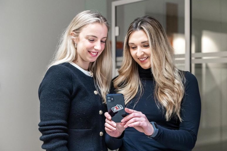 Zwei Frauen in schwarz gekleidet stehen vor einem gläsernen Büro und schauen auf ein iPhone, auf dem ein rzh-Sticker zu sehen ist, um sich über Zwangsurlaub zu informieren.