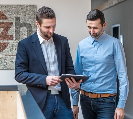 Zwei Männer, einer links mit blauem Sakko und weißem Hemd und einer rechts mit hellblauen Hemd schauen gemeinsam auf ein Tablet.