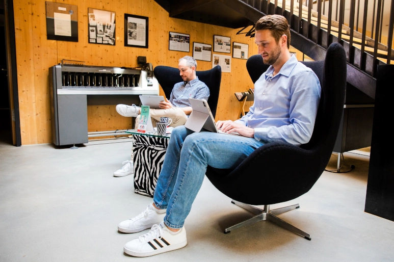 Zwei Männer, beide in hellblauem Hemd gekleidet, sitzen in jeweils einem Sessel und schauen auf ihre Laptops, die sie auf ihren Beinen abgelegt haben. Sie erkundigen sich über die Nachtarbeit.