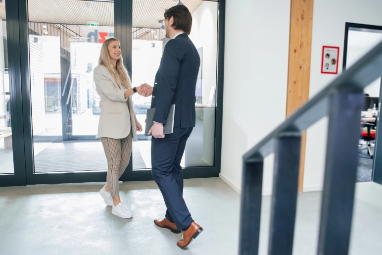 Ein Mann (Payroll Outsourcing Anbieter) und eine Frau treffen sich zu einem ersten Kundengespräch. Beide schütteln sich im Foyer die Hand.
