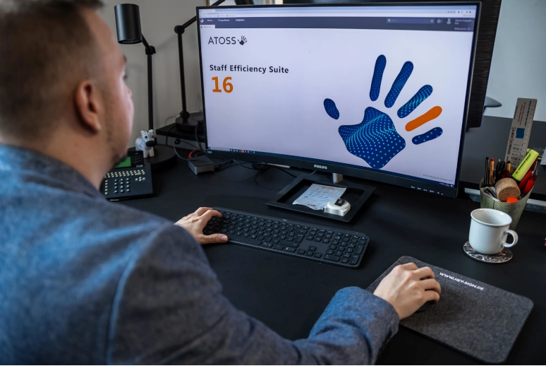 Ein Mann sitzt vor einem PC auf dem gerade ATOSS gestartet wird, um auf seinen digitalen Stundenzettel zu schauen.