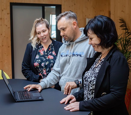 Mann in grauem Pullover steht an einem Hochtisch mit zwei Frauen in schwarzen Oberteilen. Sie schauen gemeinsam auf den Code of Conduct auf einem Laptop in ihrer Mitte.