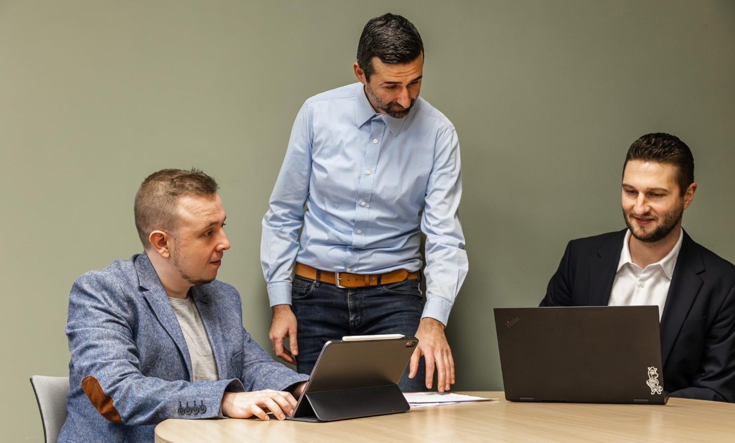Drei Männer sind in einem Büro, zwei sitzen vor ihren Laptops und ein Mann steht und erklärt den beiden Männern etwas.
