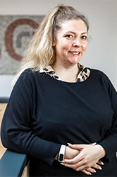 Bild von einer blonden Frau mit Pferdeschwanz und einem schwarzen Pullover, sie trägt darunter eine Bluse und man sieht ihre beige Uhr.