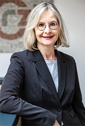 Eine Frau mit blonden Haaren und Brille, sie trägt ein graues Oberteil und einen schwarzen Blazer.