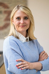 Foto von einer blonden Frau im hellblauen Pullover. Darunter trägt sie eine Bluse.