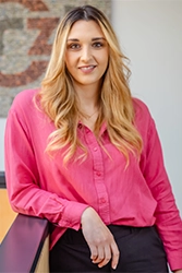 Porträtfoto von einer blonden jungen Frau am Tisch angelehnt in pinker Bluse.