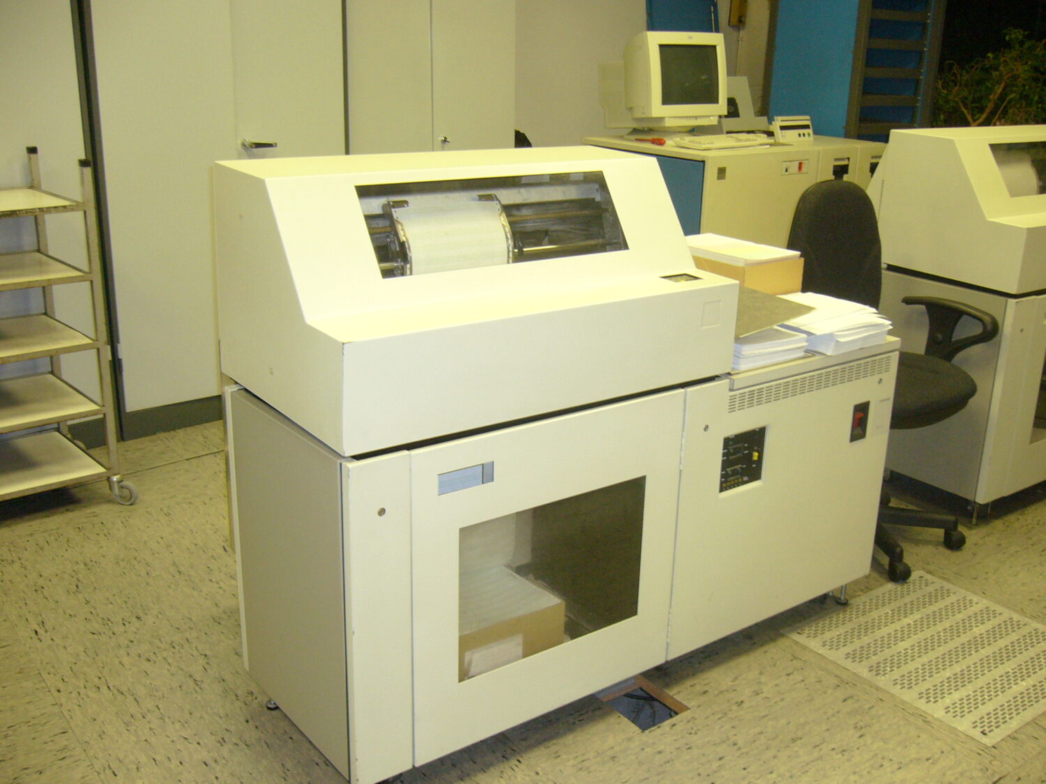 Alter Drucker mit Papier auf der Ablage und schwarzen Bürostuhl daneben stehen in einem Büro