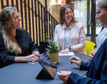 Drei Frauen stehen an einem Tisch und unterhalten sich in der Mitte steht ein Tablet.
