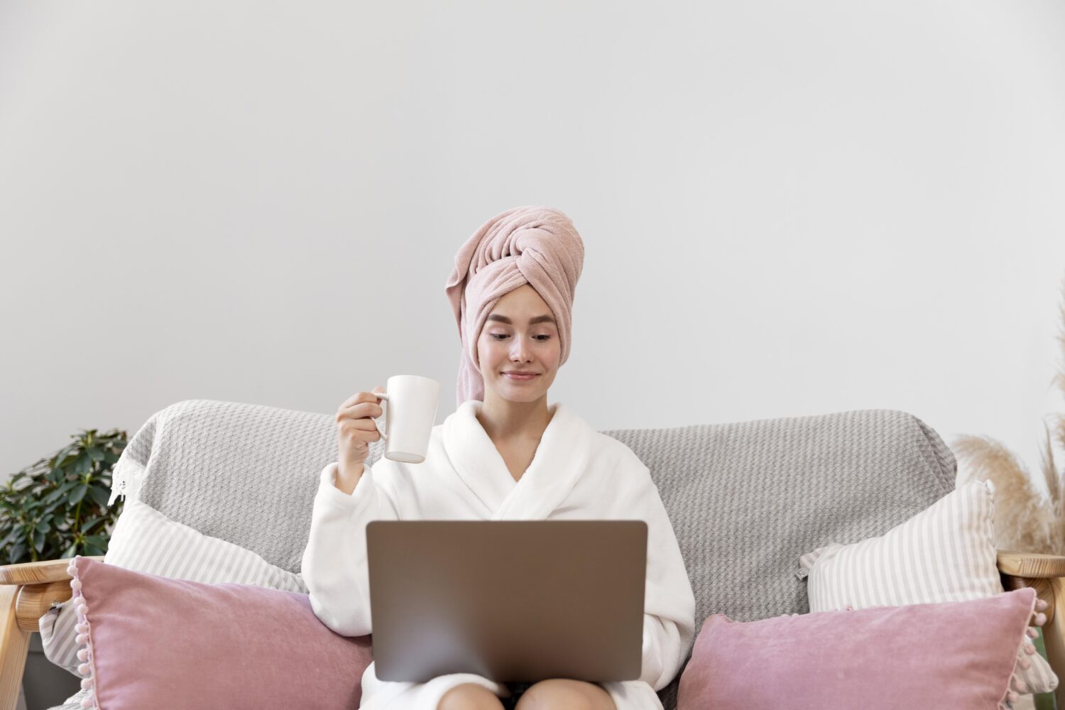 Eine Frau sitzt auf ihrem Sofa zuhause und arbeitet im Bademantel am Laptop mit einer Tasse in der Hand.