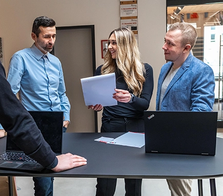 Ein Mann in hellblauem Hemd, ein Mann in hellblauem Blazer und eine Frau in schwarzer Kleidung stehen an einem Hochtisch. Die Frau hält einen Block in die Höhe. Der Mann in dem Blazer hat die Zeitwirtschaft auf seinem Laptop offen.