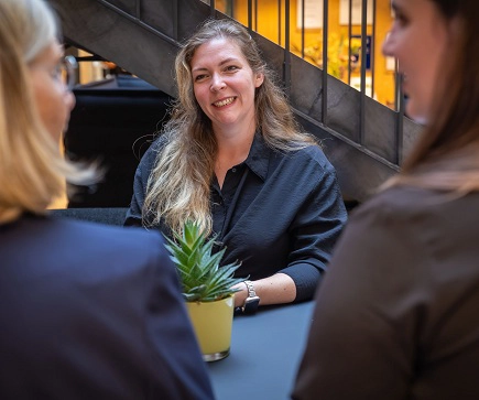 Eine blonde Frau im schwarzen Oberteil unterhält sich.